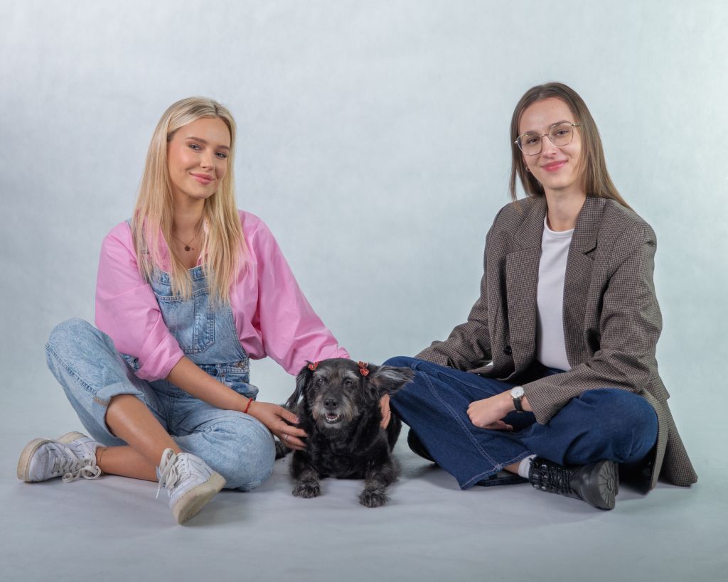 Na zdjęciu widoczne są dwie osoby siedzące na podłodze na jednolitym, jasnym tle. Między nimi znajduje się mały czarny pies. Obie osoby mają swobodną, nieformalną postawę i delikatnie głaszczą zwierzę. Scena ma spokojny, przyjazny charakter.